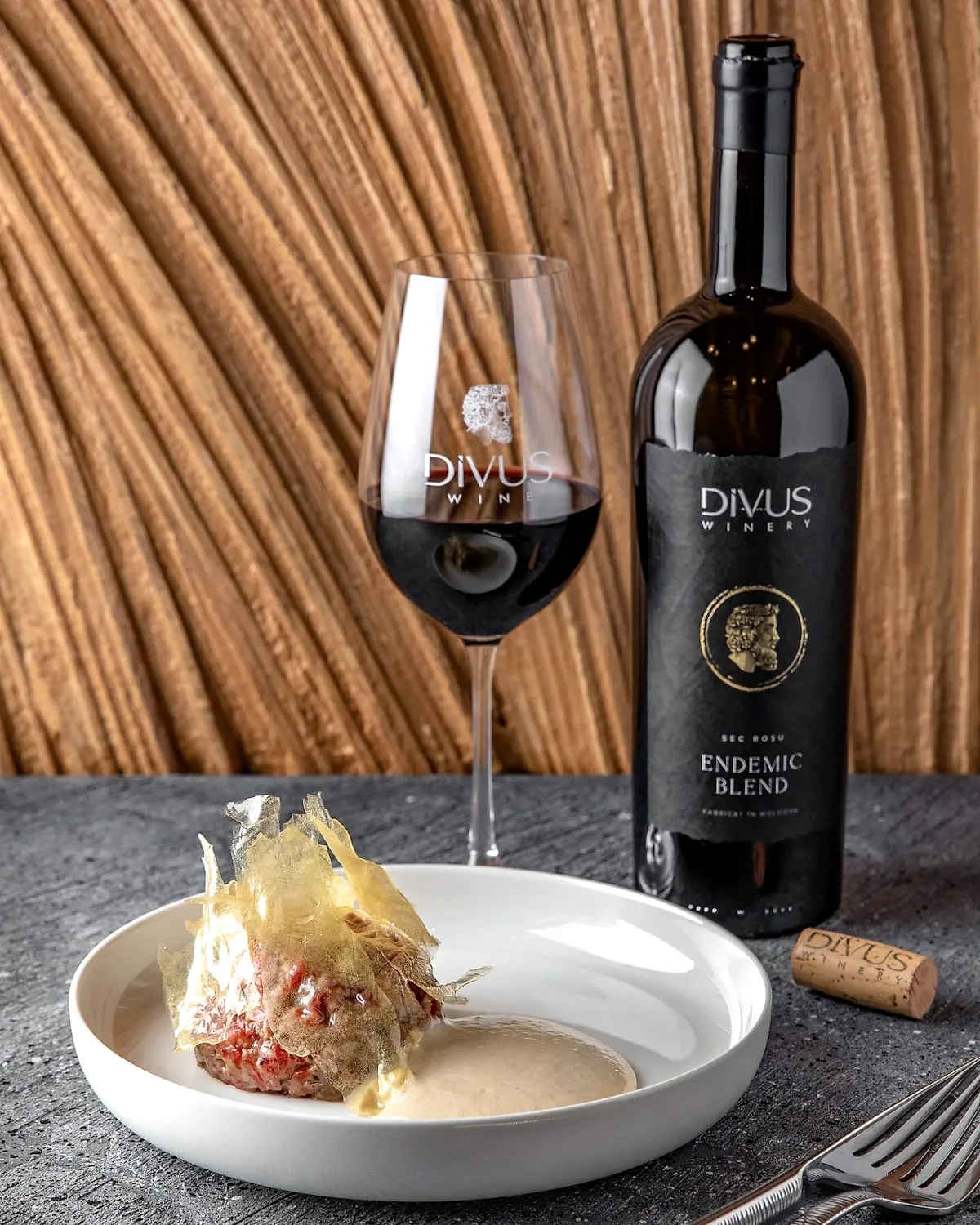 A bottle of Divus and a glass of red wine beside a dessert